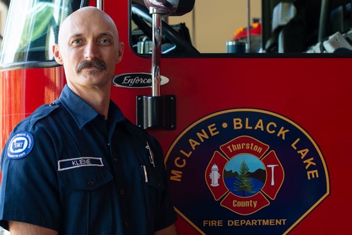 The image features a firefighter in uniform posing next to a red fire truck with the McLane-Black Lake Fire Department emblem.