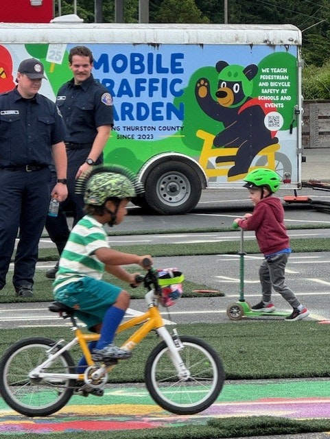 A child on a bike and another on a scooter near police officers and a colorful Mobile Traffic Garden truck promoting safety.