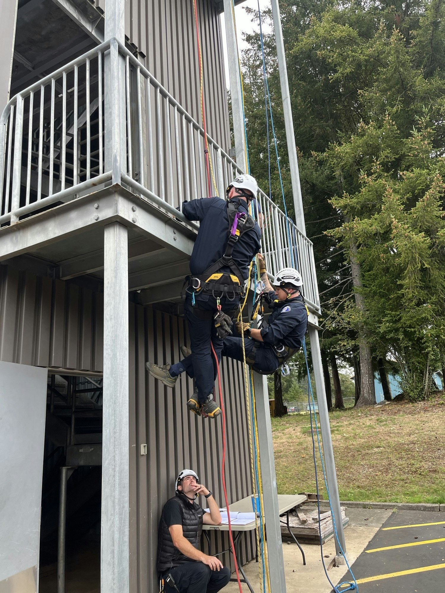 The image shows a group practicing rope rescue techniques on a building, with safety gear and harnesses involved.