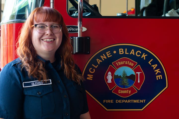 A smiling person in a fire department uniform stands next to a red fire truck with a logo for McLane Black Lake Fire Department.
