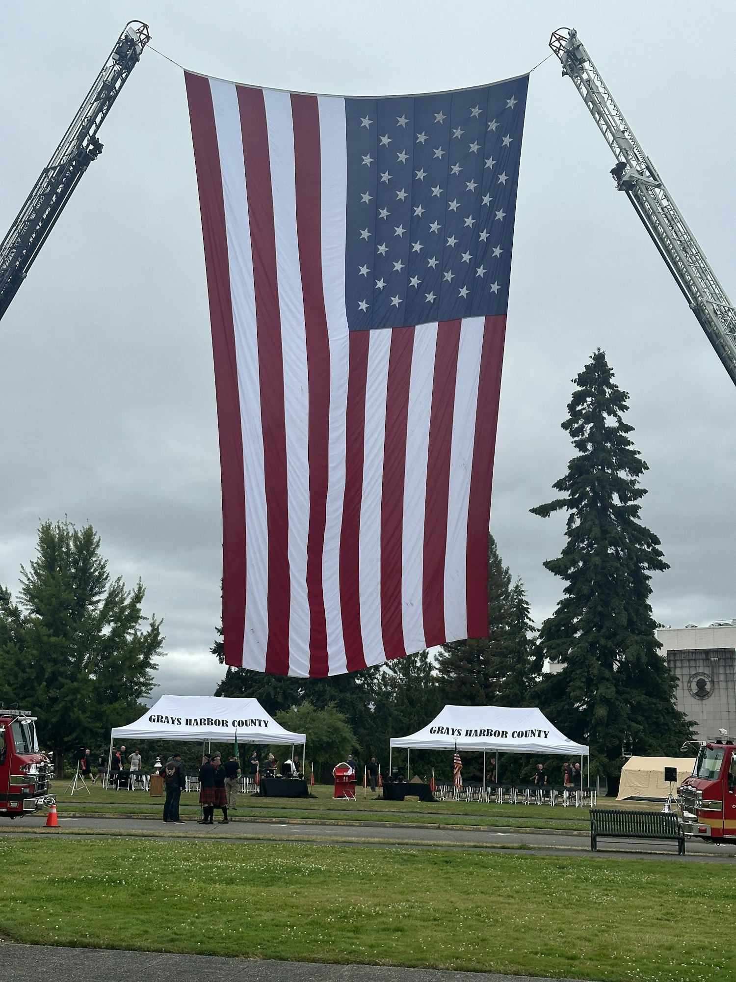 A large American flag is displayed prominently, suspended by fire truck ladders at an outdoor event in Grays Harbor County.