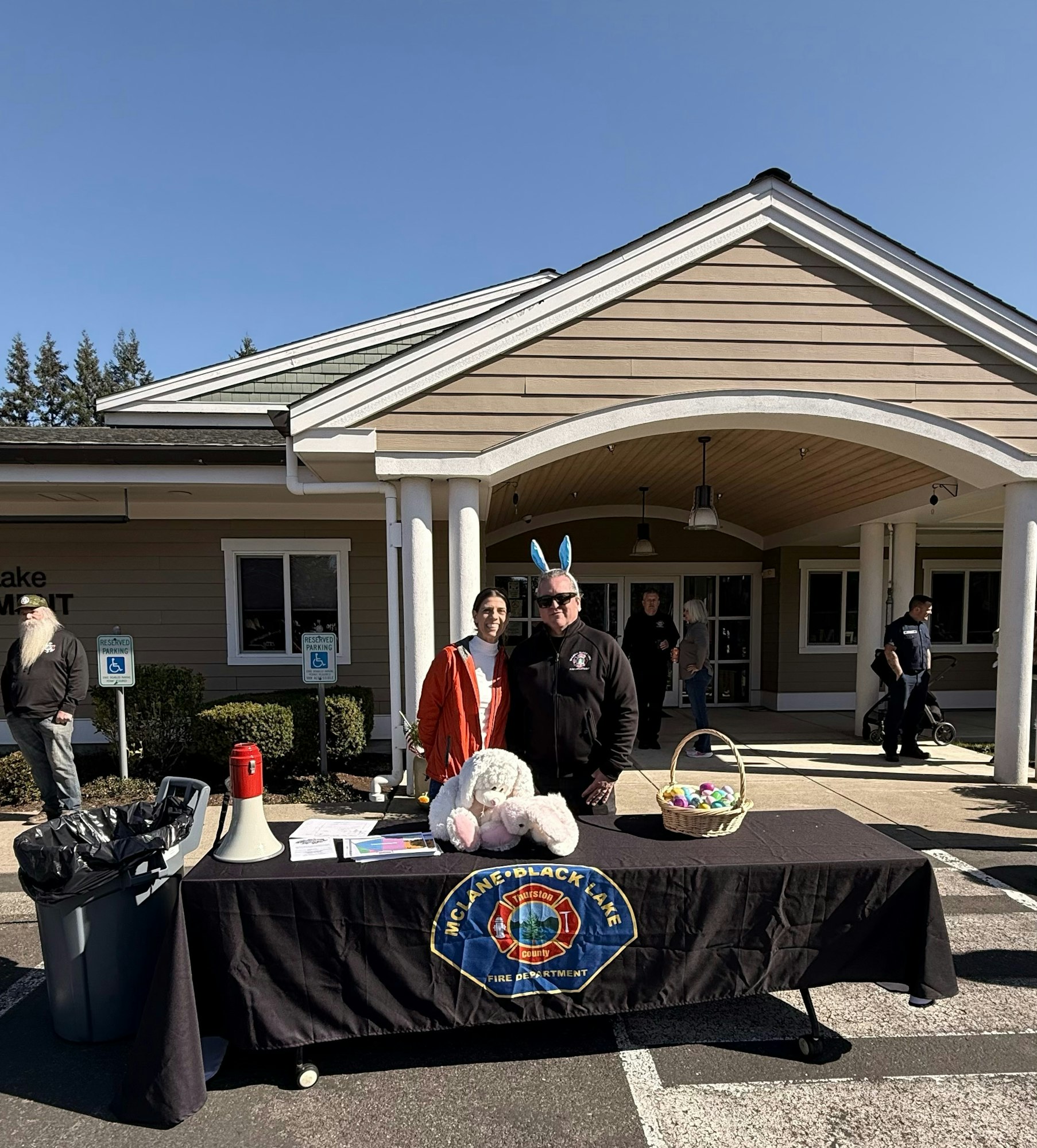 A table with decorations, a giant bunny, two volunteers, and a basket of eggs at a community event outside a building.