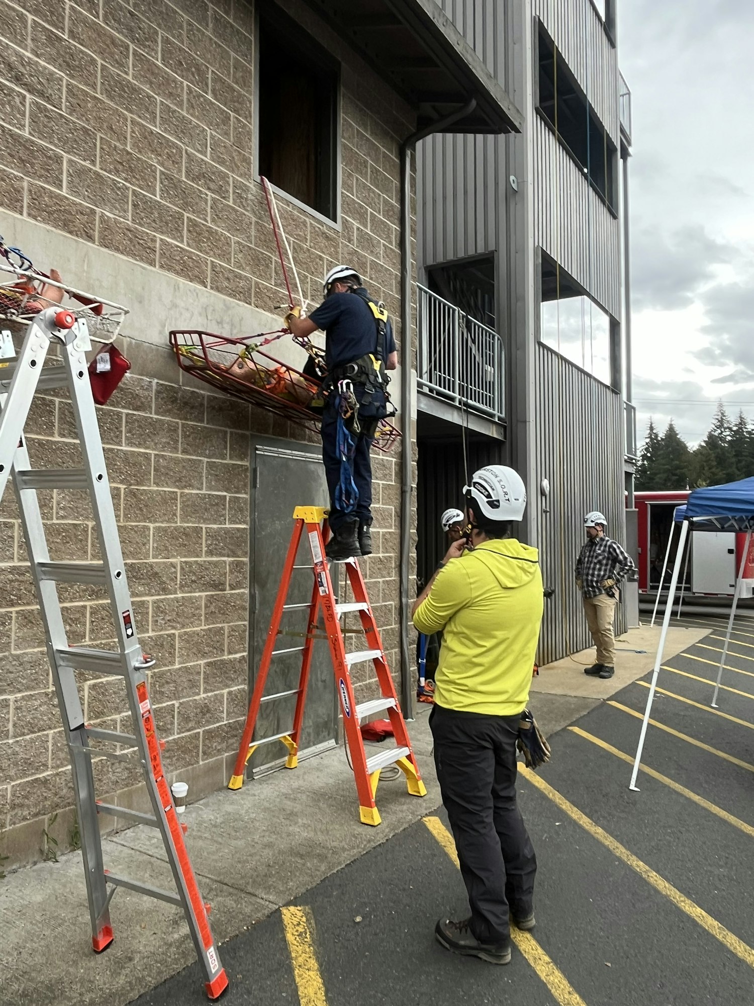 A worker in safety gear is using a scaffold to work on a building while another person observes nearby. Ladders are also present.