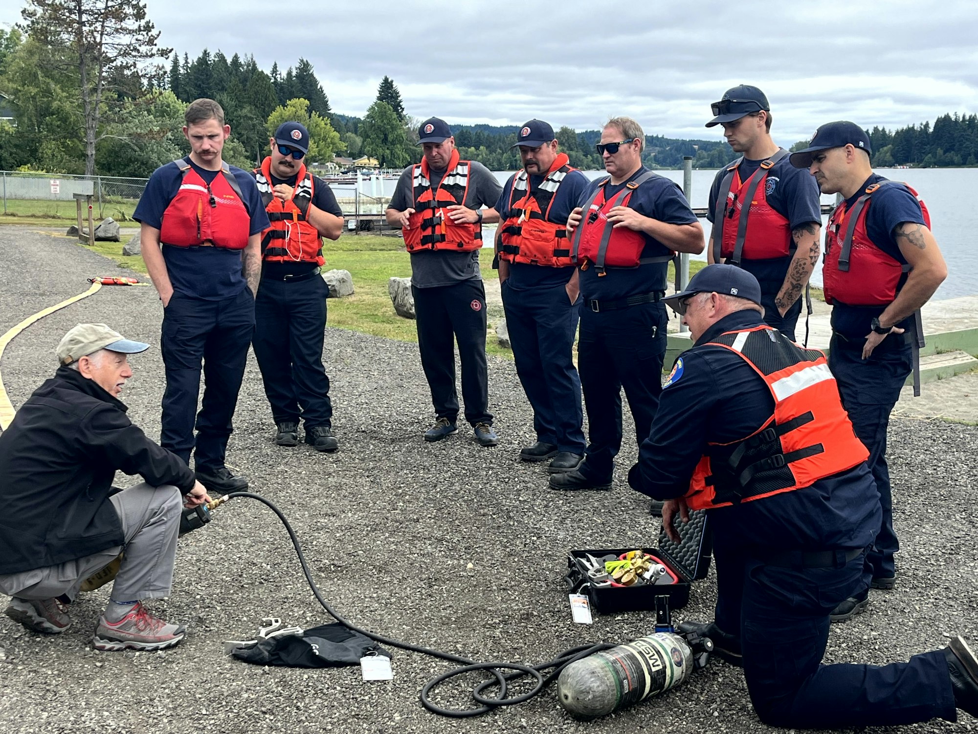 A group of men in life vests listens to an instructor, possibly during a water safety or rescue training session.