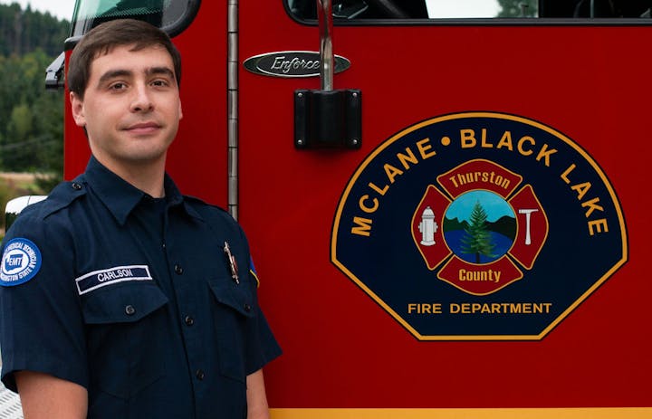 A person in a uniform stands beside a fire truck labeled "MCLANE BLACK LAKE FIRE DEPARTMENT."