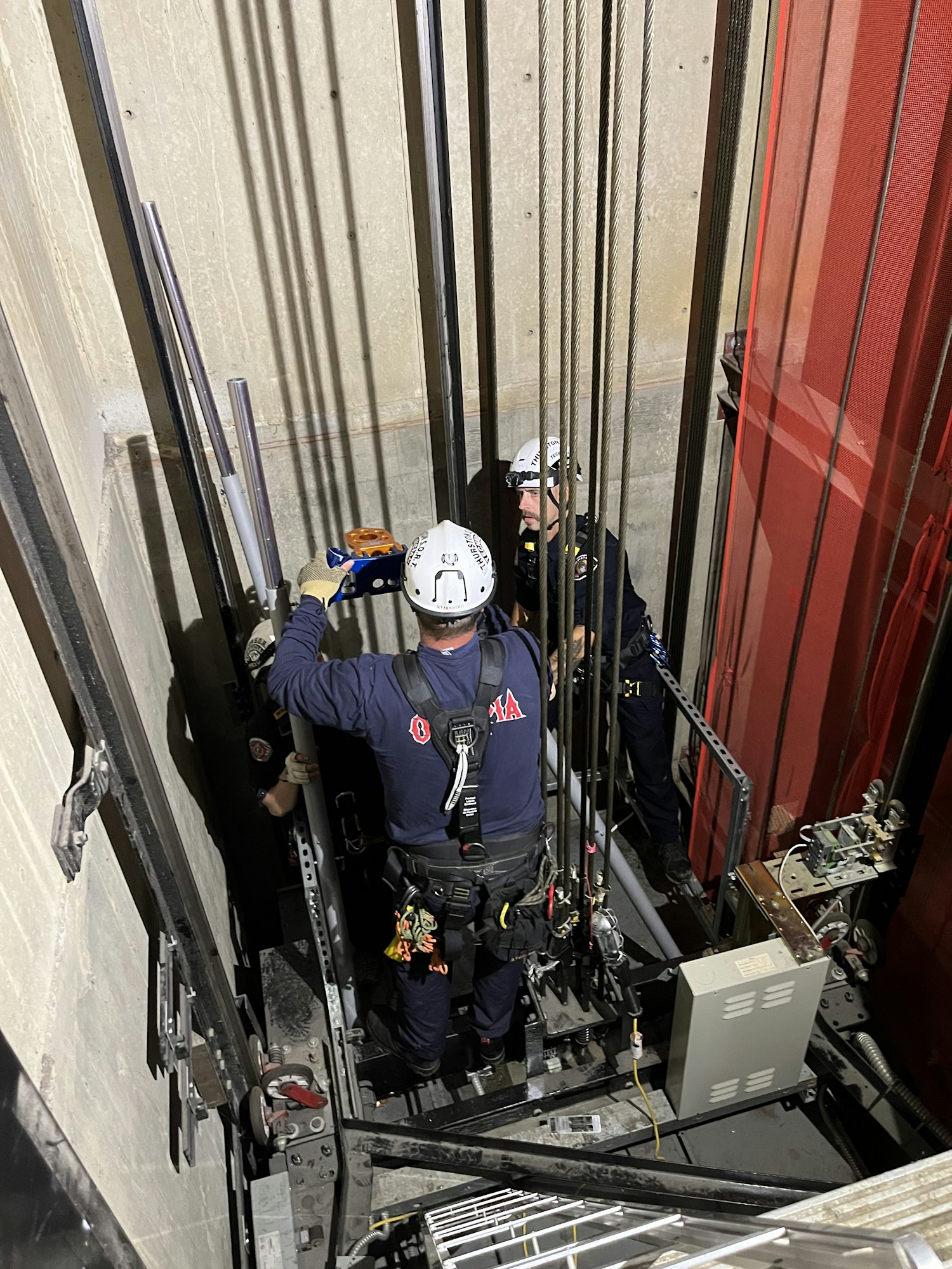 Two workers in helmets are performing maintenance inside an elevator shaft, surrounded by cables and machinery.