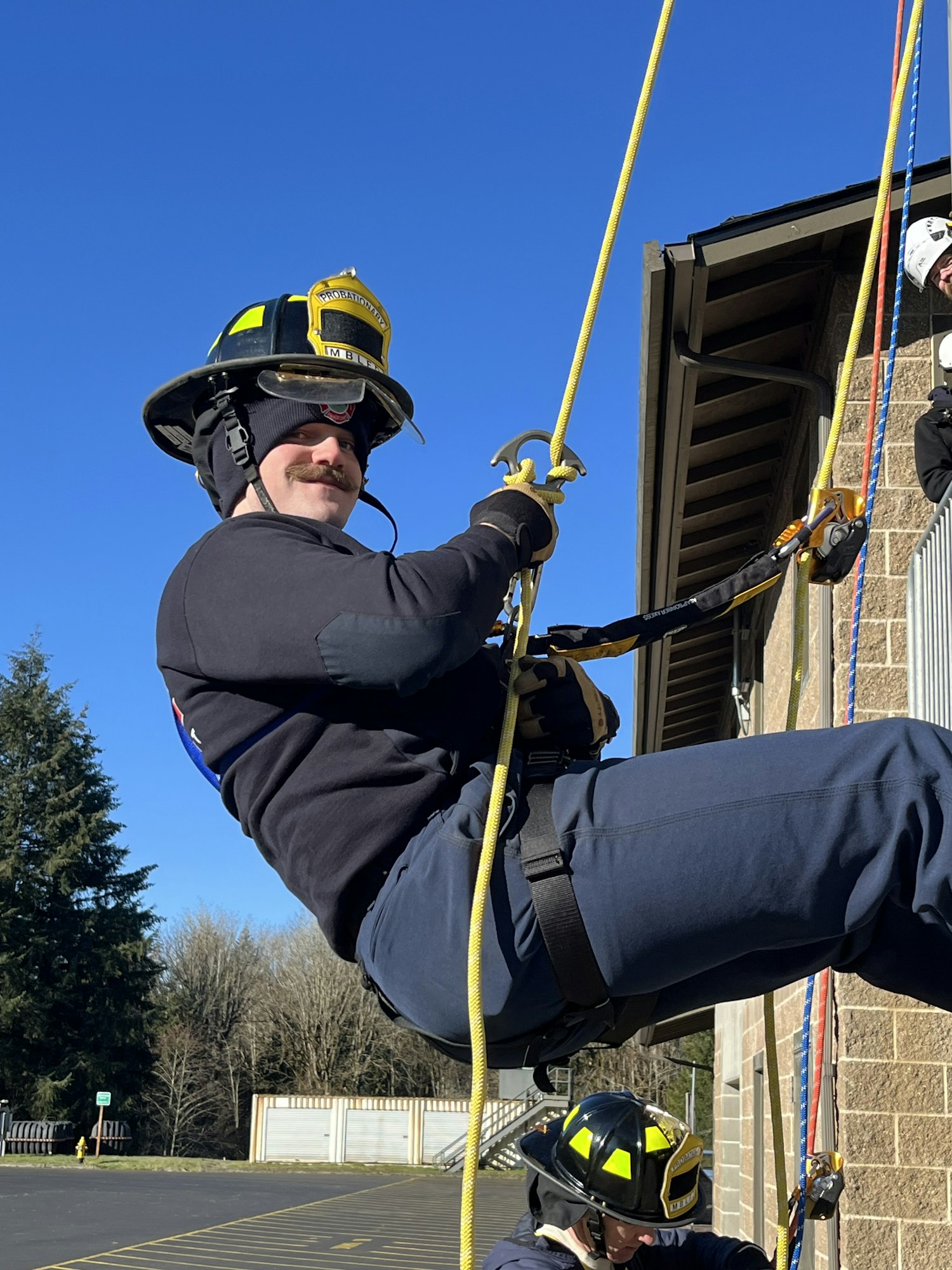 A firefighter is rappelling down using ropes, wearing protective gear and a helmet, with clear blue skies in the background.