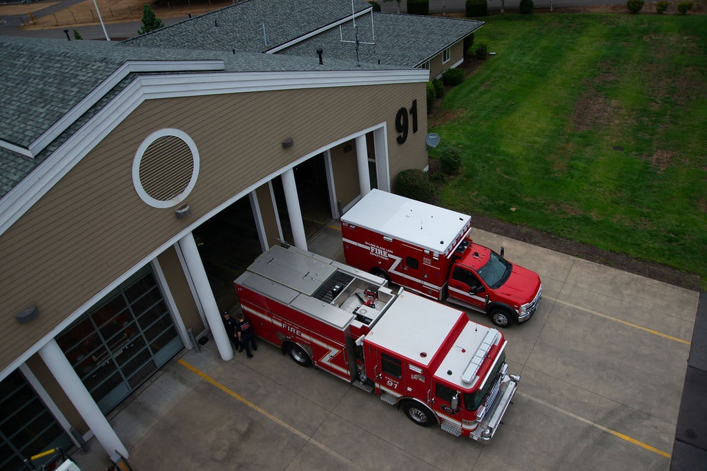 The image shows a fire station with two emergency vehicles parked outside, surrounded by a grassy area.