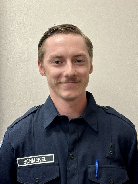 A portrait of a smiling individual in a dark blue uniform with a nameplate, standing against a light-colored background.