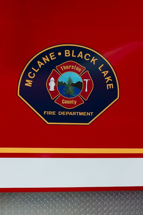 McLane Black Lake Fire Department logo with Thurston County badge, hydrant, tree, and axe on a red backdrop.