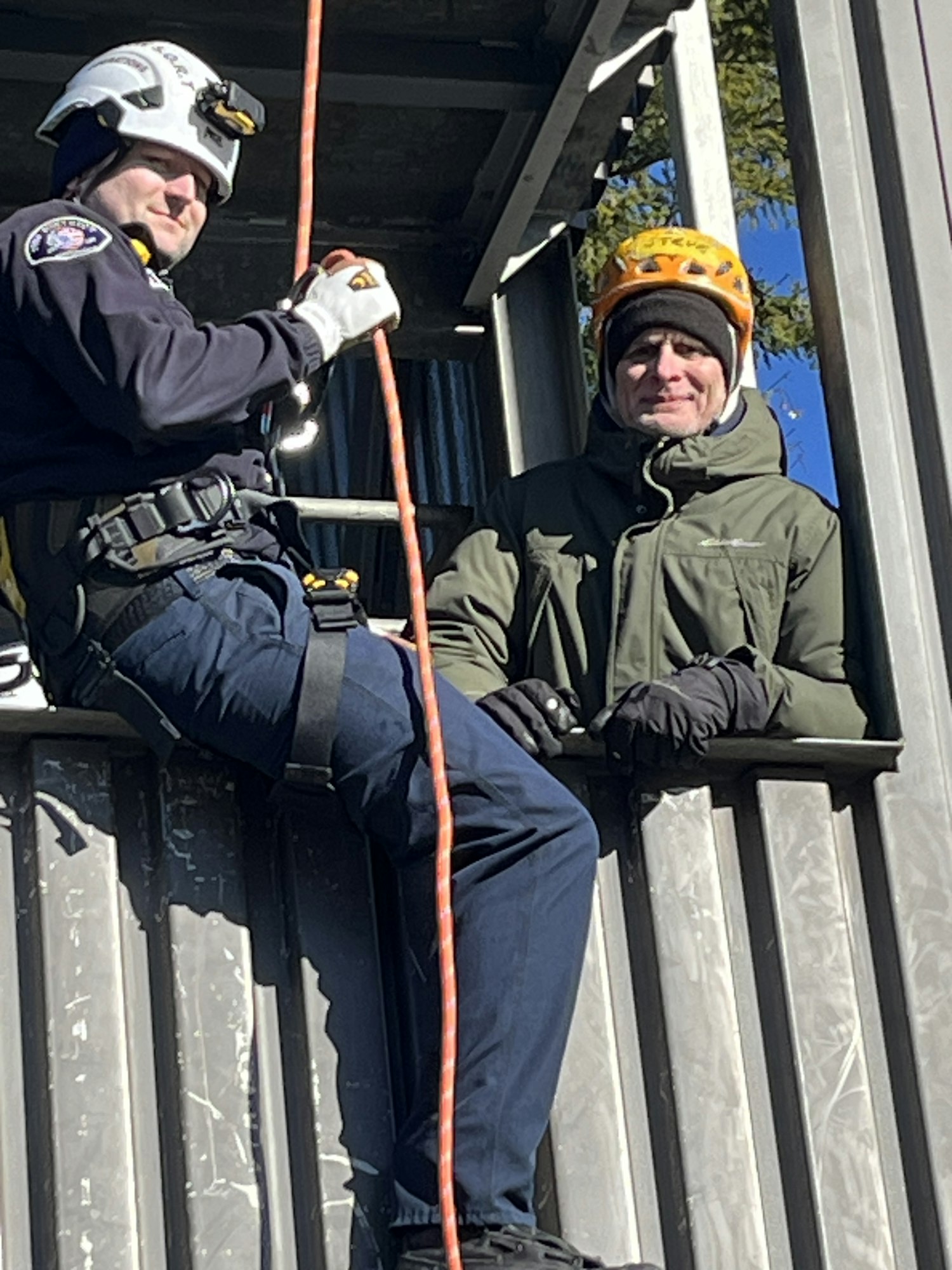 The image shows two individuals in helmets, one climbing and the other positioned at a height, using climbing gear and ropes.