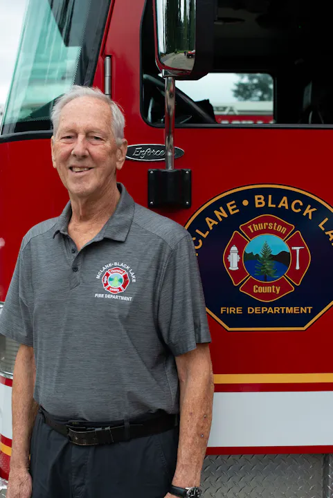 A smiling older man stands in front of a red fire truck, wearing a fire department shirt, with a logo visible behind him.