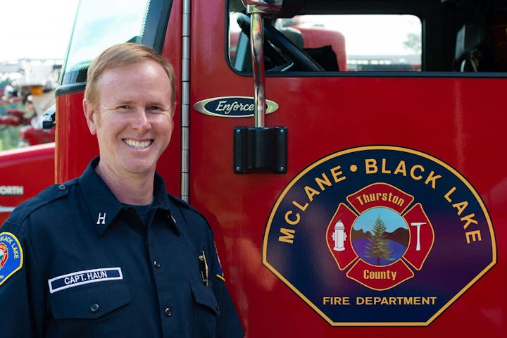 A smiling firefighter standing in front of a red McLane-Black Lake fire truck.