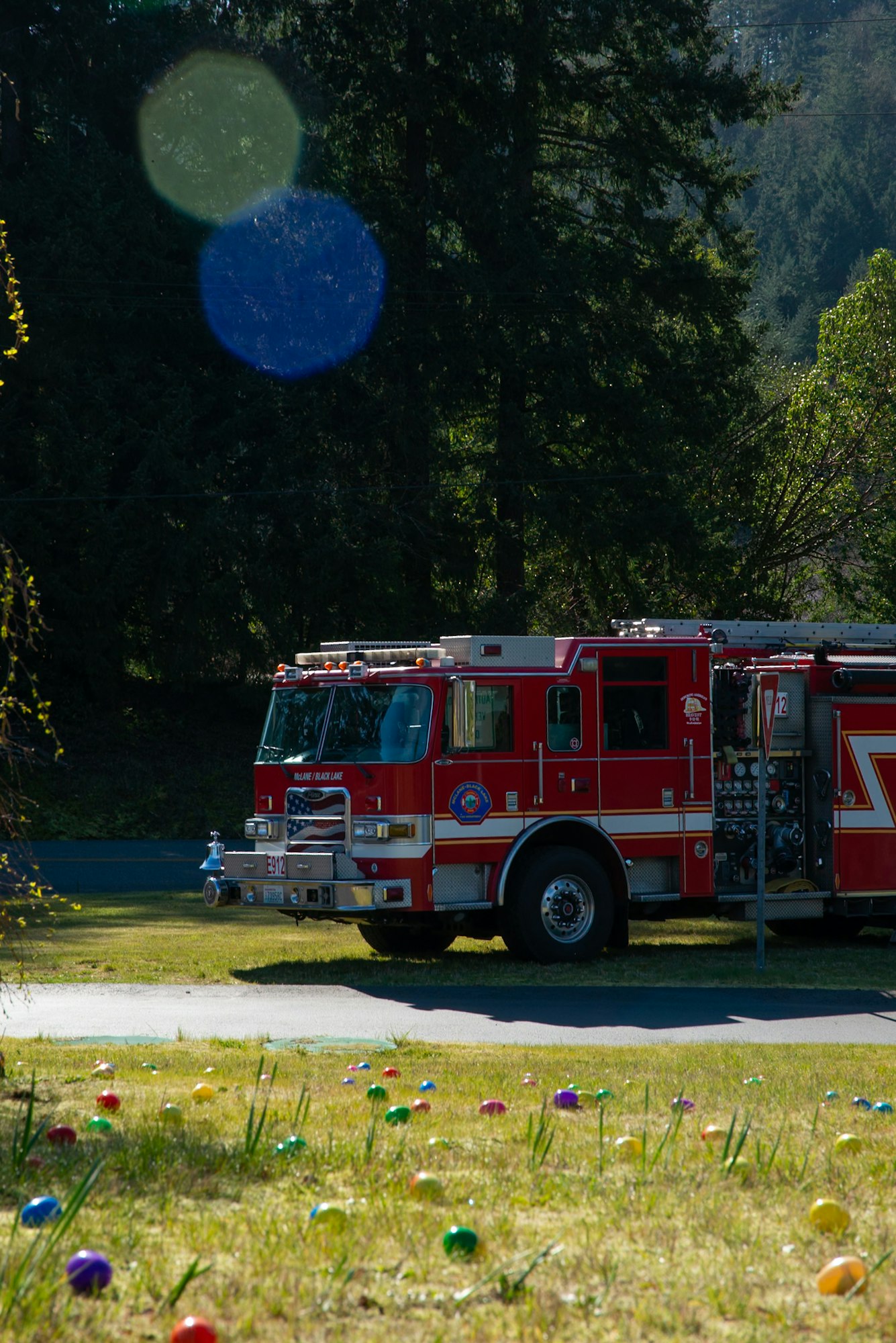 A red fire truck parked near colorful decorative eggs on grass, surrounded by trees and a clear sky.