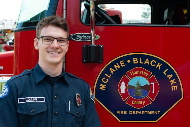 A person in a fire department uniform stands next to a red fire truck with the McLane Black Lake Fire Department logo.