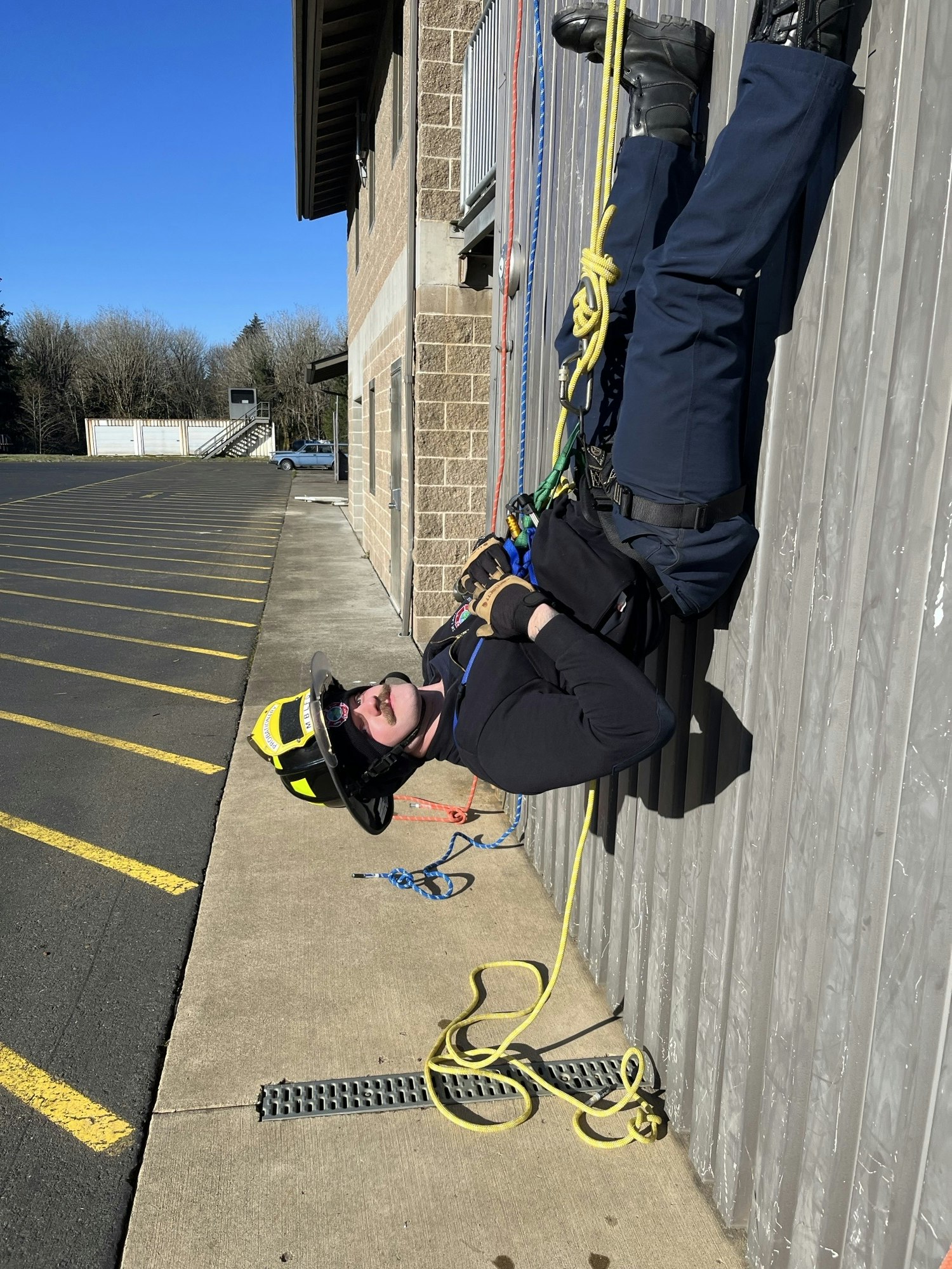 A person in safety gear is roped and hanging upside down from a building, practicing rescue techniques outdoors.