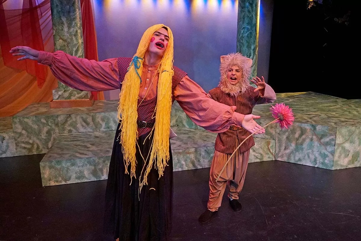 Two people in theatrical costumes performing on stage, one with a long yellow wig, the other holding a pink flower.