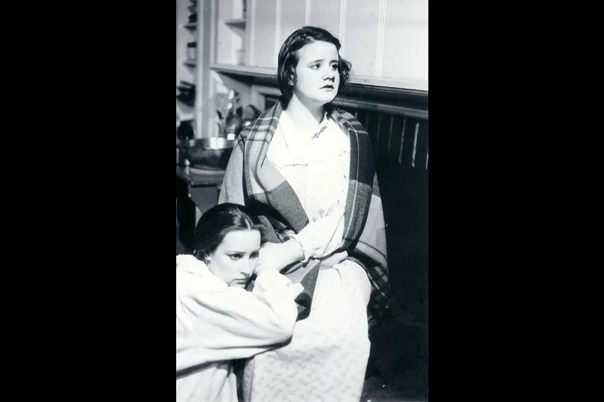 Two women in a black-and-white photo, one sitting and wrapped in a blanket, the other sitting close to her.