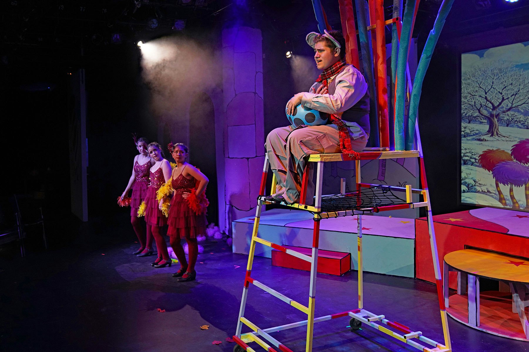 A theatrical performance scene featuring a performer on a ladder and three dancers in vibrant costumes on stage.