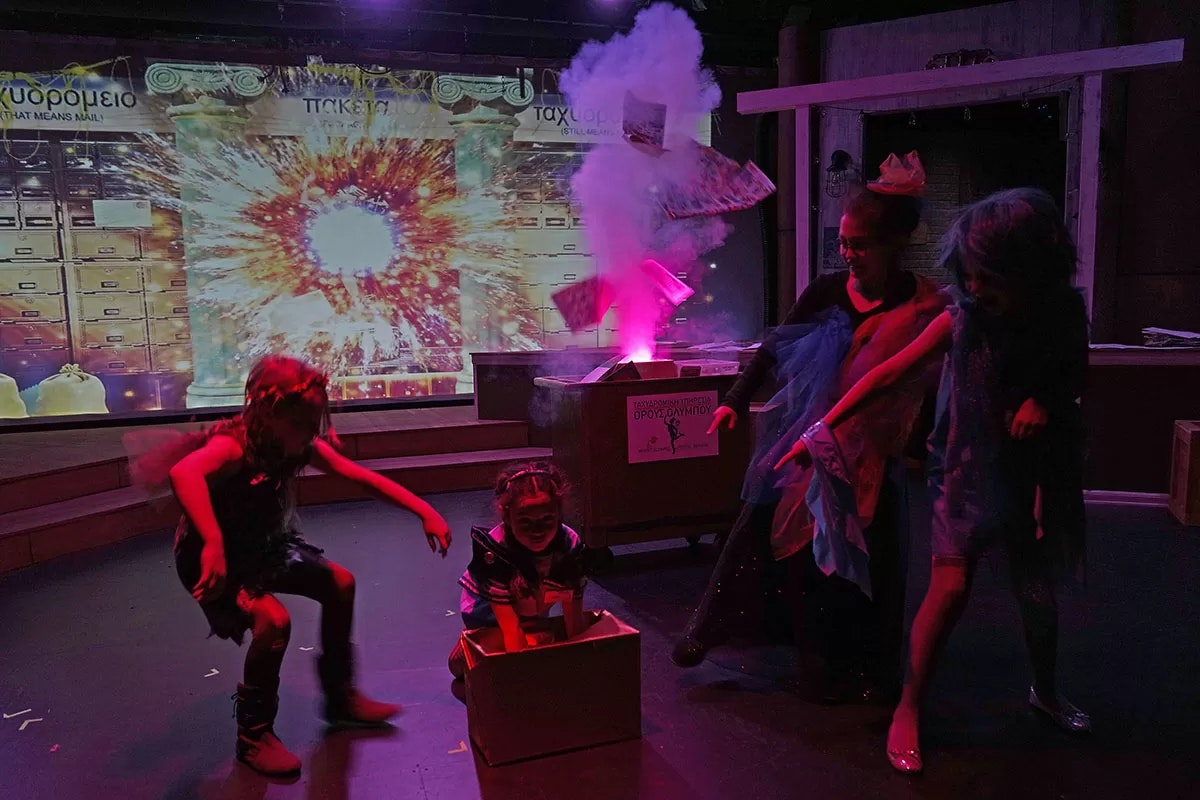 Four children in costume perform on stage; a backdrop shows mailboxes and an explosion. Smoke and papers rise from a box.