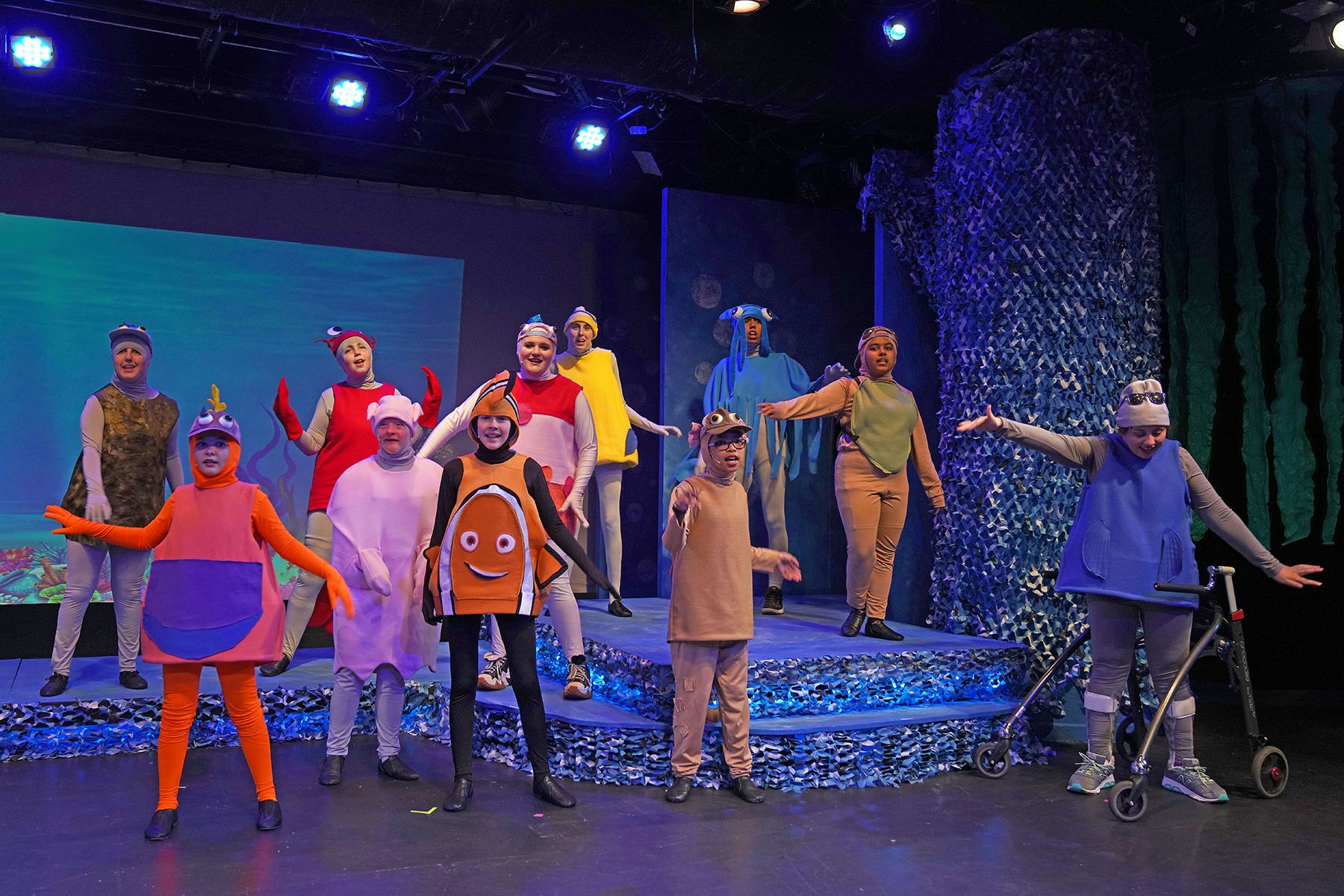 A group of performers in colorful sea-themed costumes on stage, with oceanic set design and lighting.