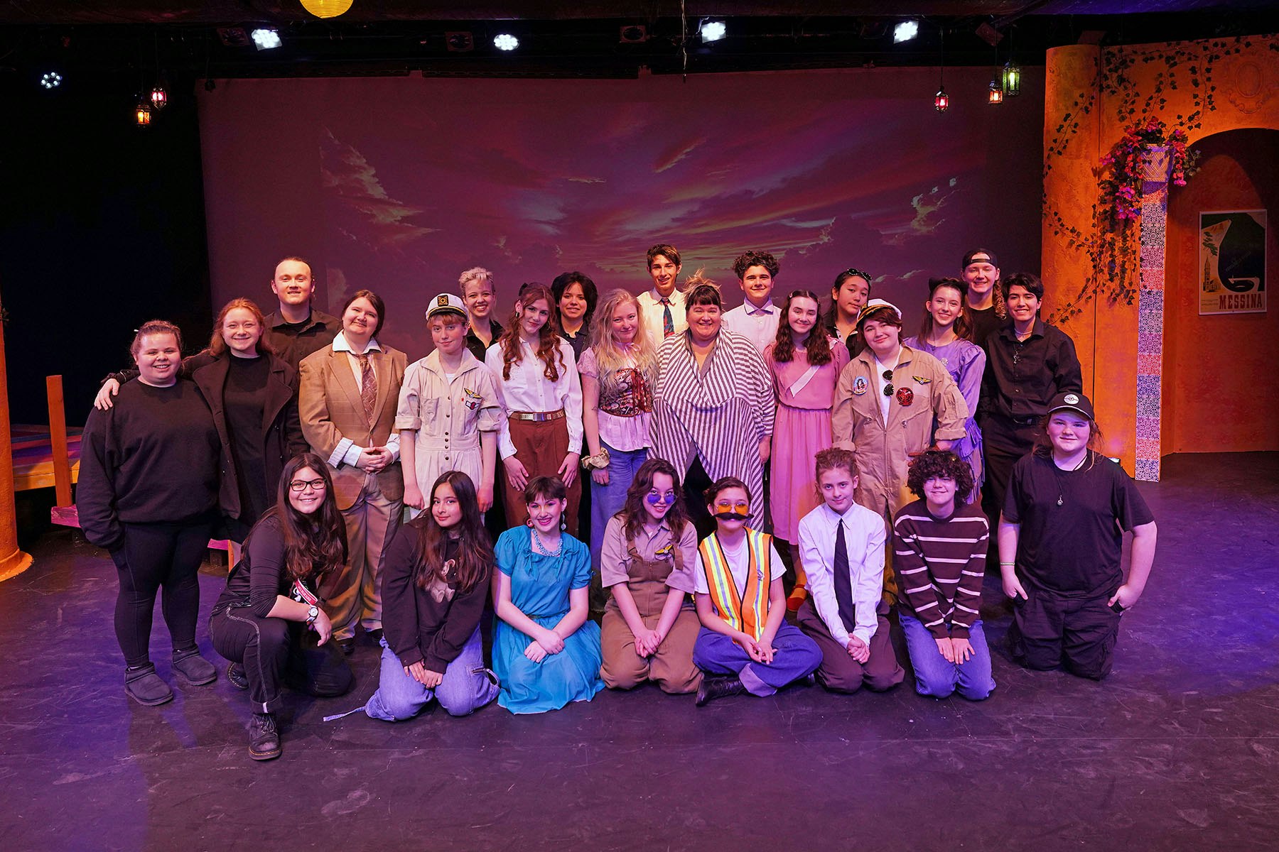 A group of about 25 people in various costumes posing on a stage with a decorative backdrop.