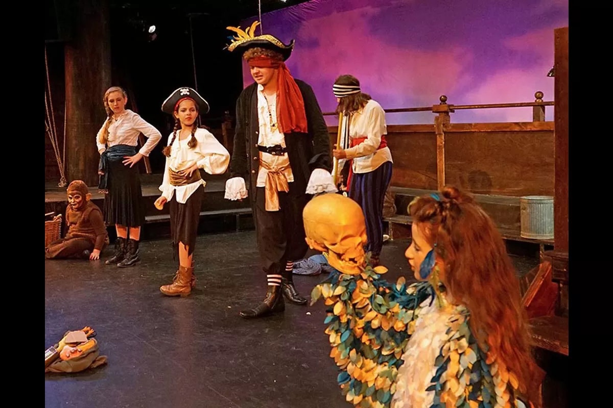 A theatrical scene with people in pirate costumes, one holding a skull, on a stage with a purple backdrop.