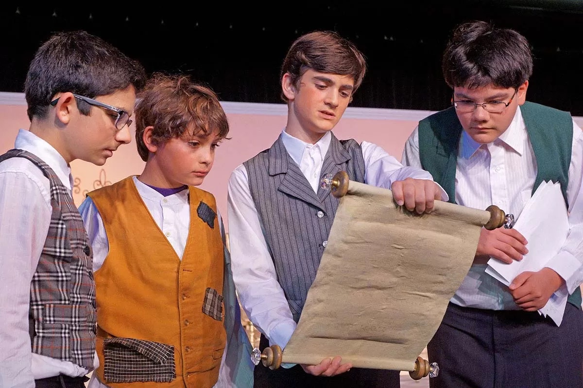 Four boys in period costumes are closely examining a scroll.