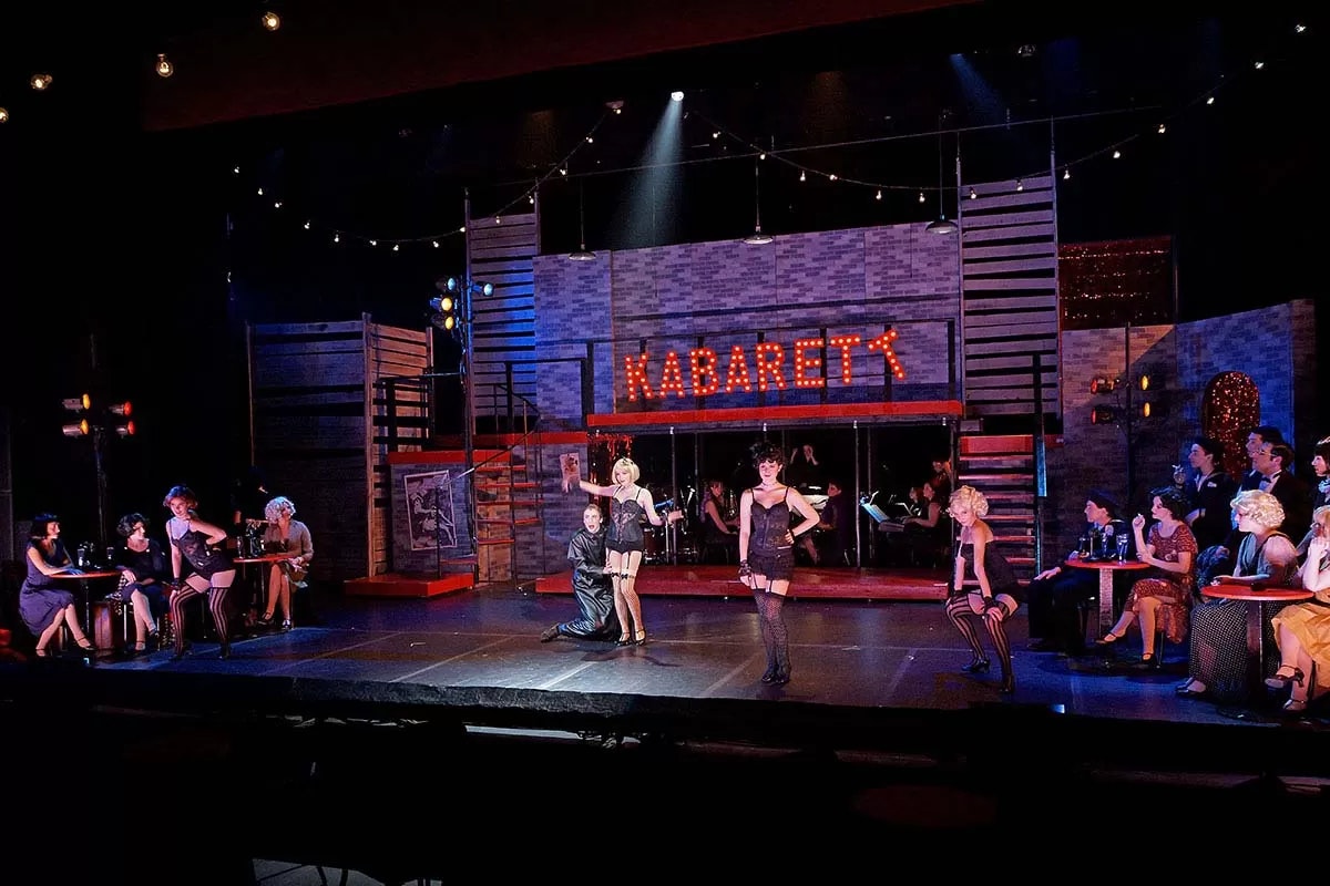 A theatrical performance on stage with performers in front of a "KABARET" sign, surrounded by seated audience members.
