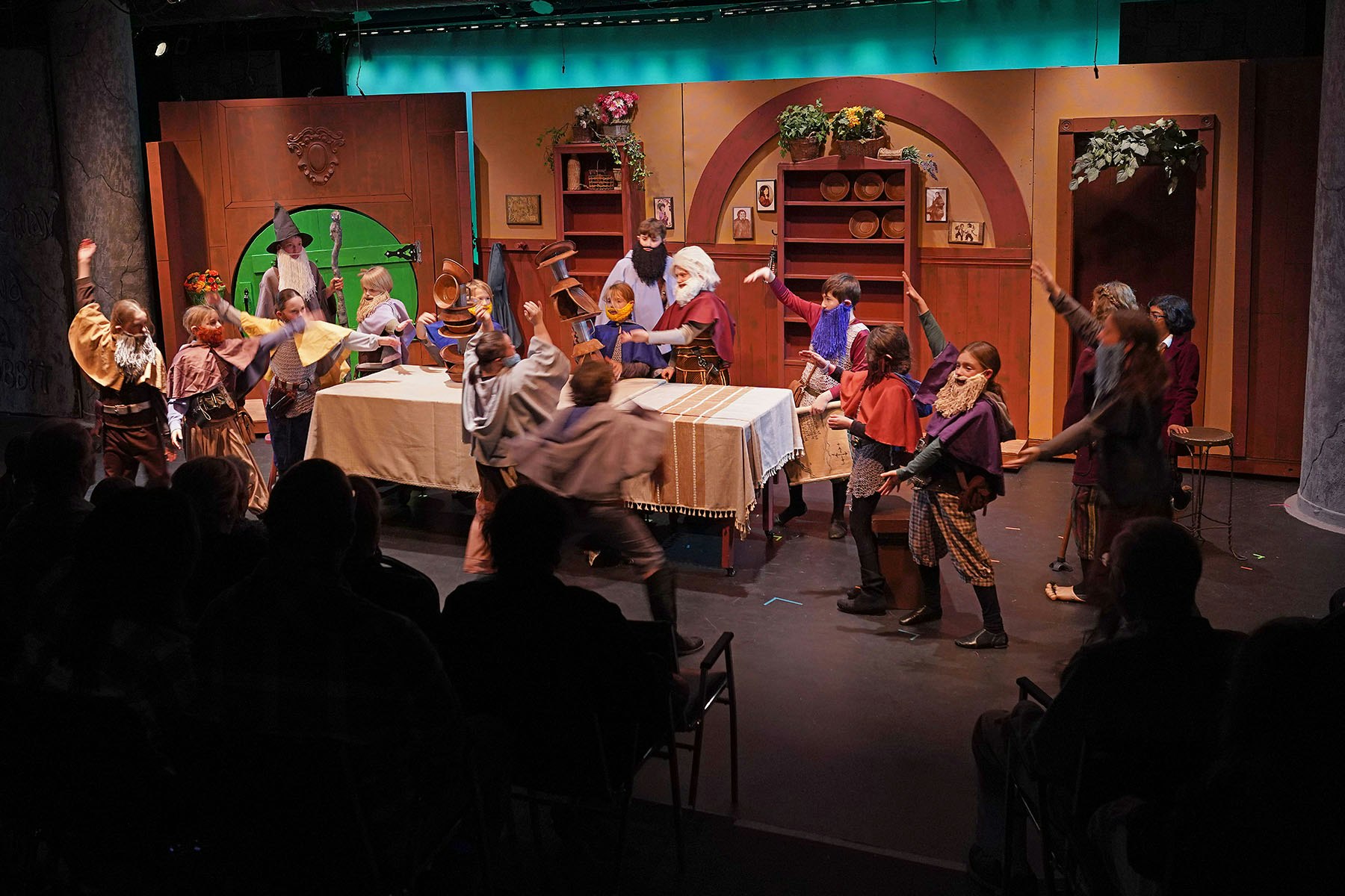 A theater scene with actors in medieval costumes performing energetically on stage, with a visible audience in the foreground.