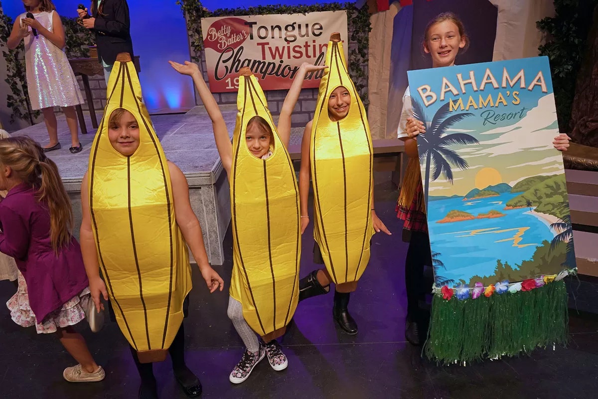 Children in banana costumes with a tropical resort sign at an event or performance.