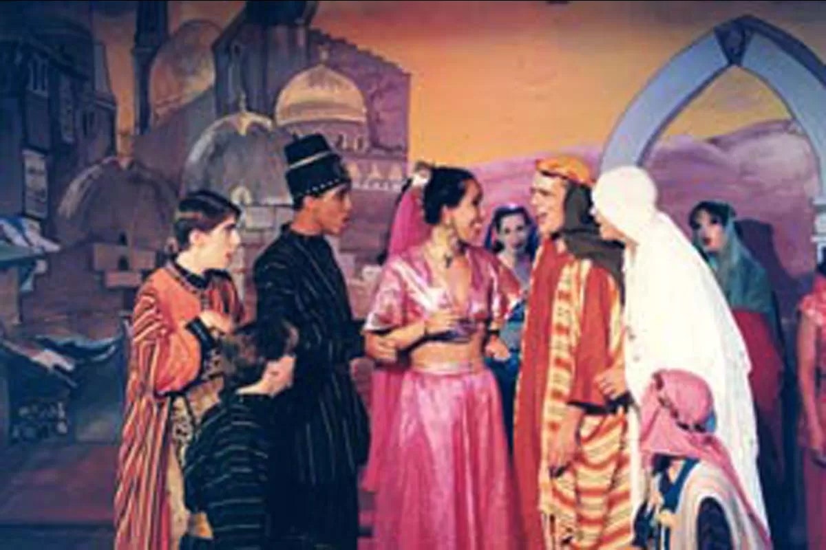 A group of people in colorful costumes perform on a stage with an ornate backdrop resembling a historical or fantasy setting.