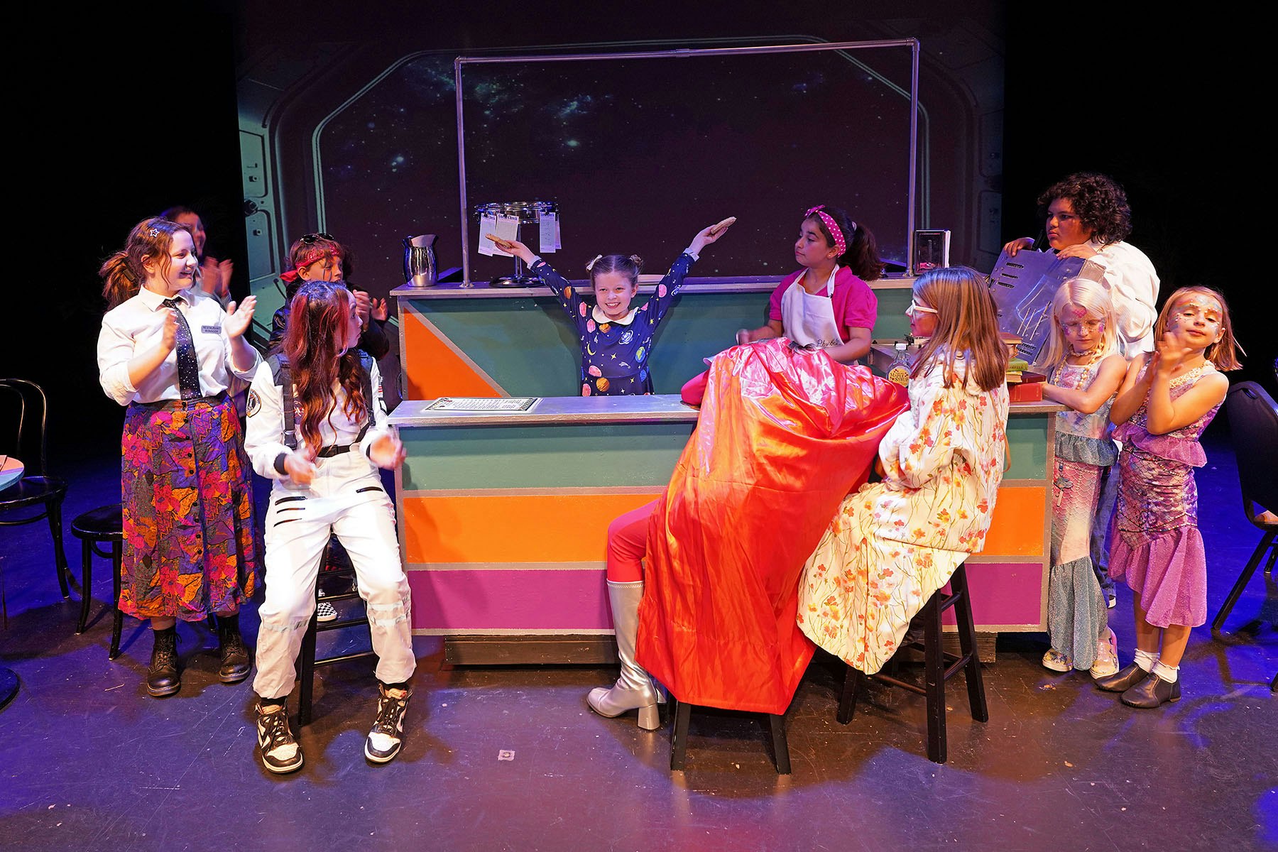 A group of children in colorful costumes perform on stage, with one child in the center raising their arms, creating a lively scene.