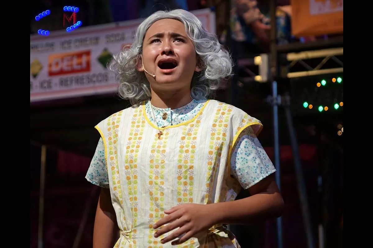 A person with gray hair, wearing a floral apron, appears to be singing or acting on stage.