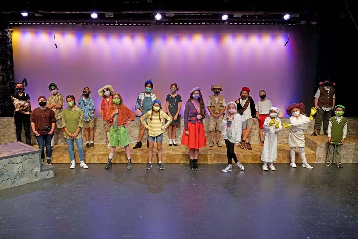 A group of children in colorful costumes and masks stand on stage under purple lighting.