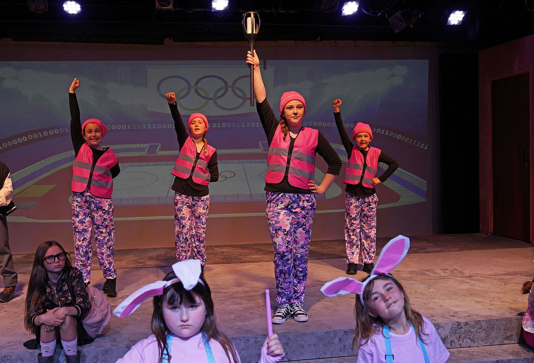 A performance featuring kids in colorful outfits, posing energetically on stage with an Olympic-themed backdrop.