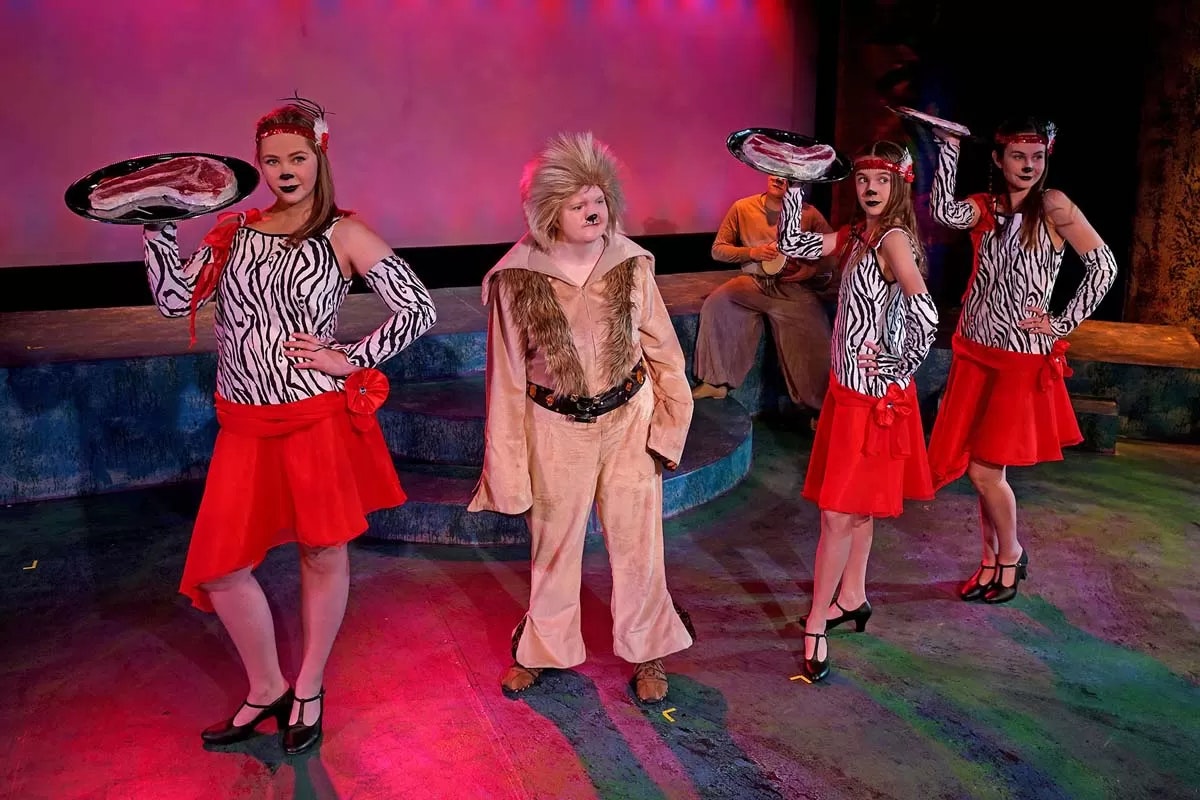 Performers in colorful animal-themed costumes with trays, likely on stage during a play or musical.
