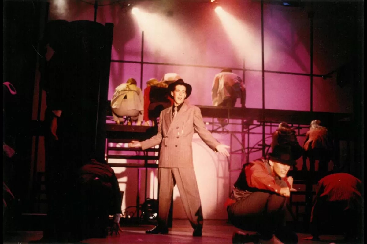 A man in a suit and hat performs on stage, surrounded by dancers in a dramatic, colorful setting.