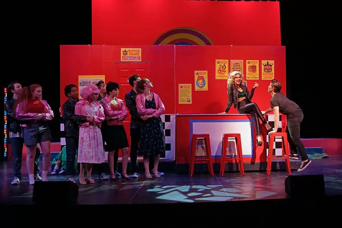 A stage performance with a retro diner backdrop, actors in 1950s-style outfits, and a woman sitting on a counter.