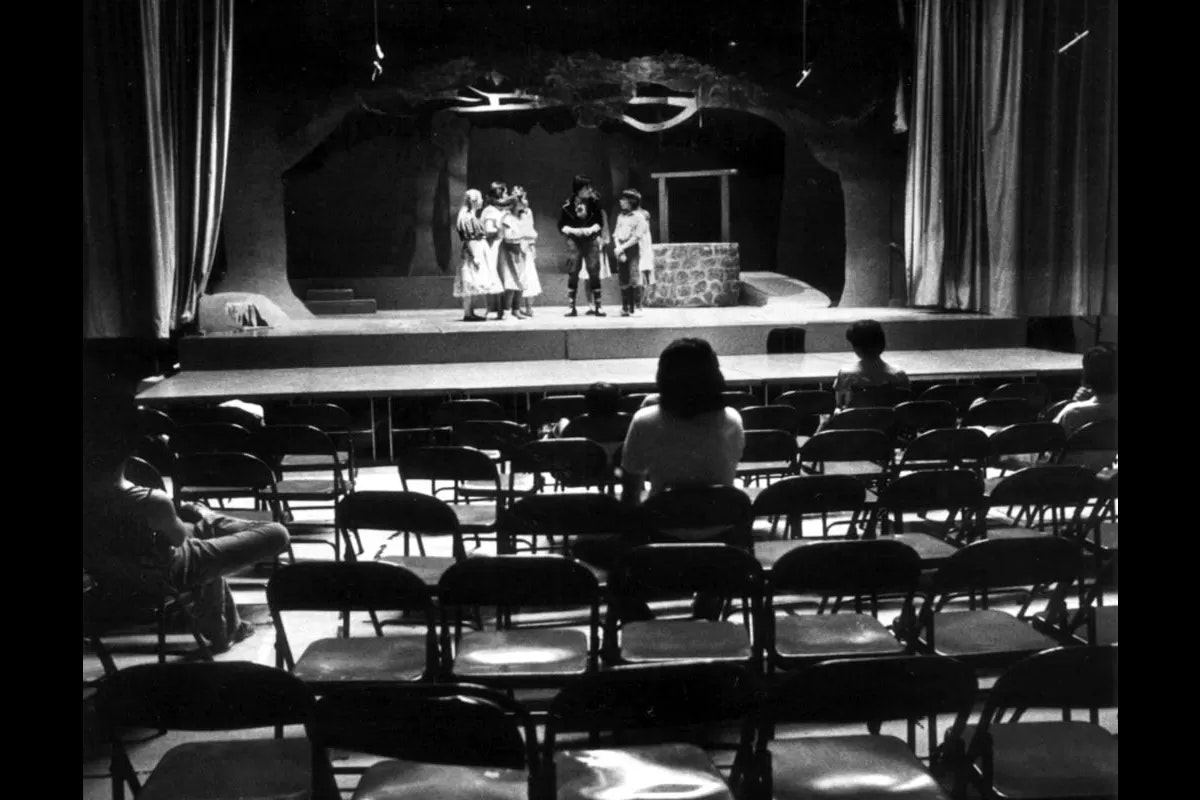 A stage with a few people performing, and an audience of a few in a room with rows of empty chairs.