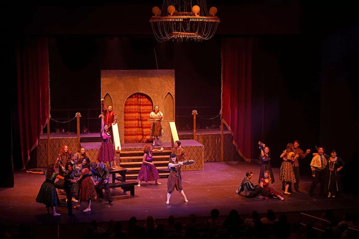 Stage play with actors in costumes, a set resembling a castle, and a chandelier above, performing a scene with expressive gestures.