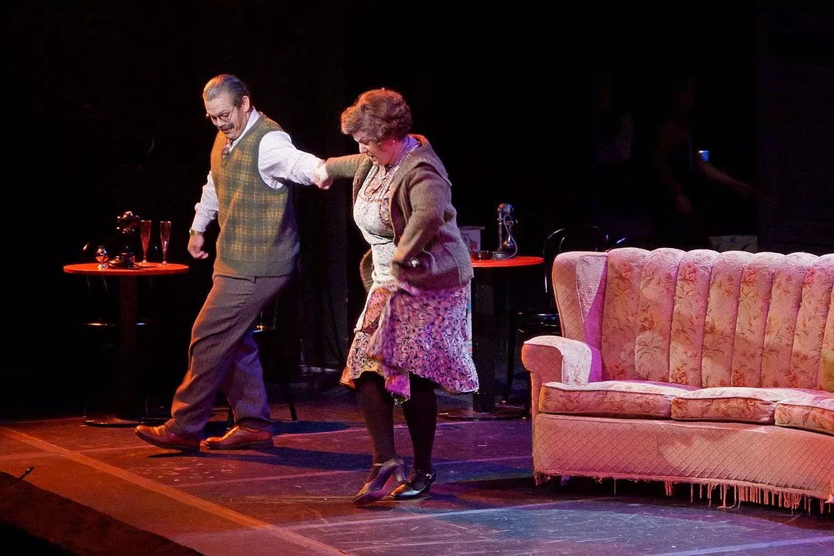 Two actors perform a lively scene on stage, with a sofa and tables as part of the set.