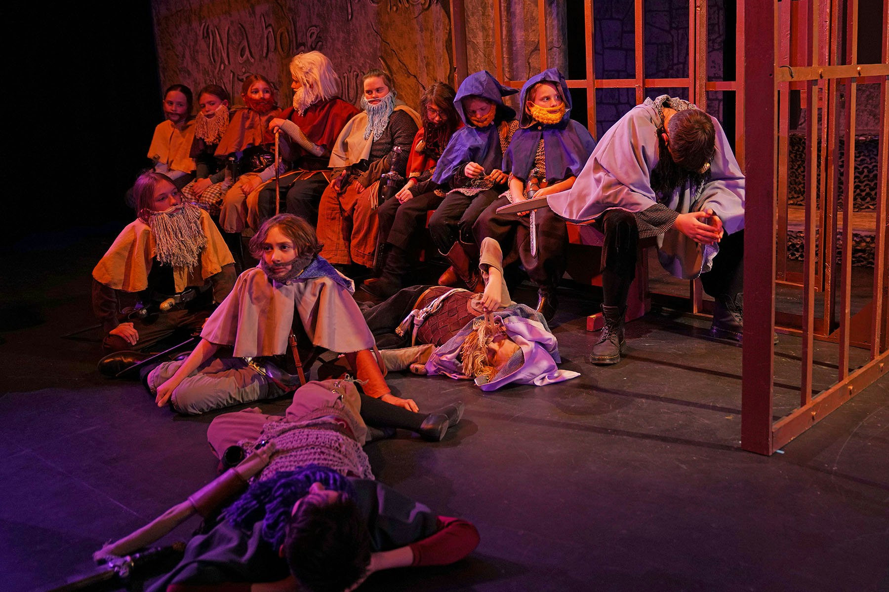 A group of people in medieval-themed costumes, some with fake beards, seated or lying on a stage, possibly during a theater performance.