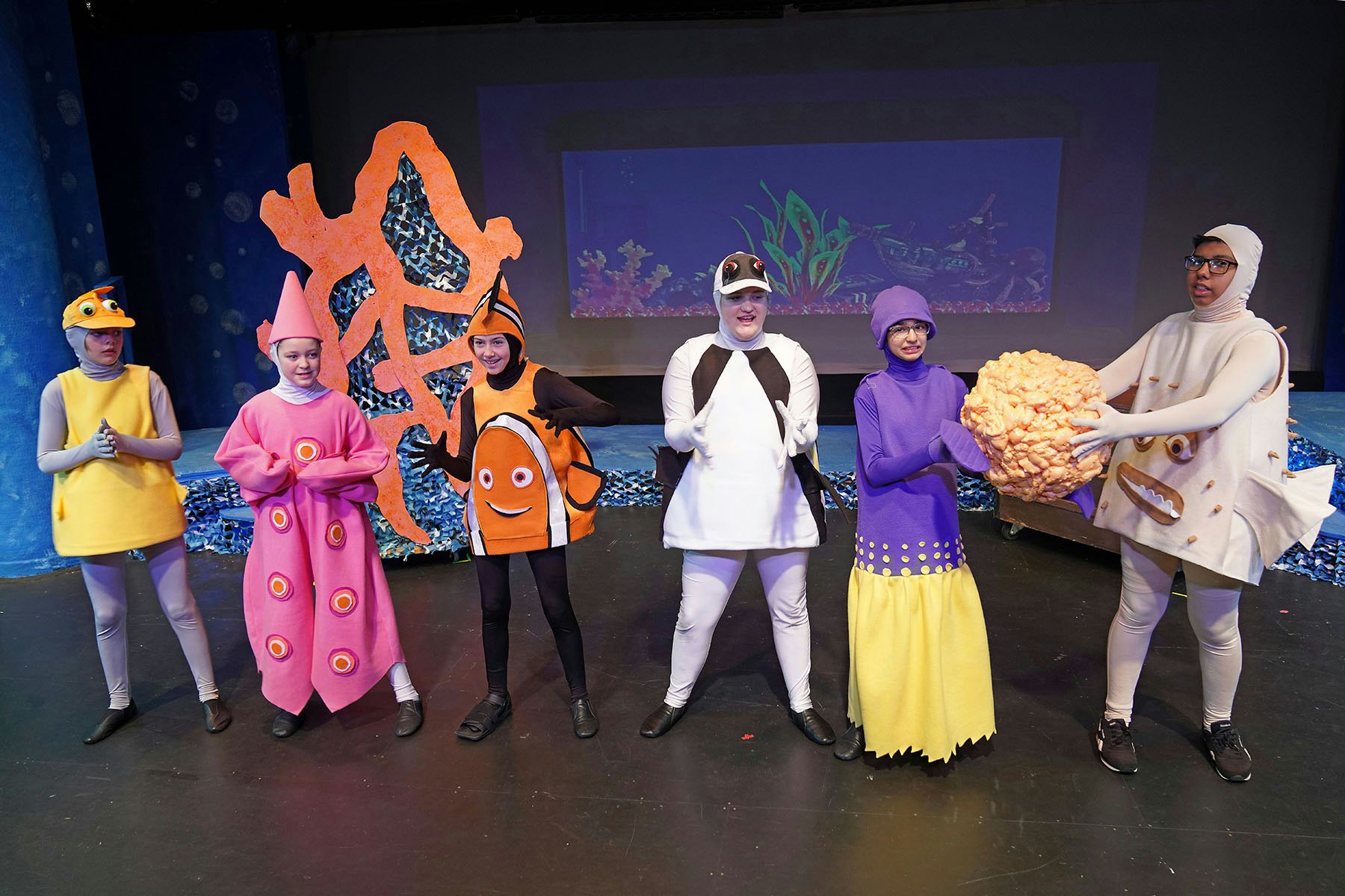 People in colorful sea animal costumes on a stage with an ocean-themed backdrop.