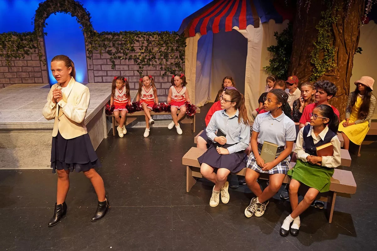 A group of children on stage, one speaking, others dressed as students or cheerleaders, with a tent and decorations in the background.
