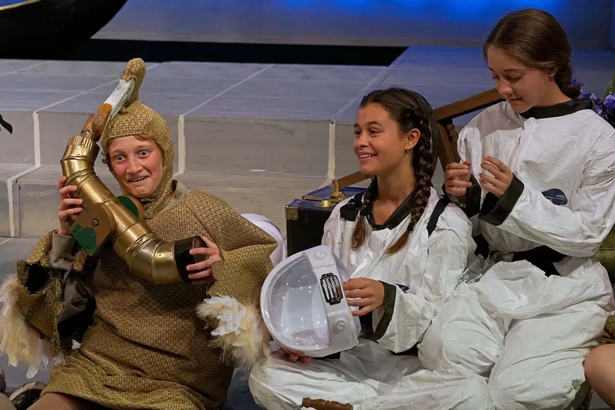 Three people in costumes: one in a bird suit with a mechanical wing, two in astronaut outfits holding a helmet, sitting on stage.