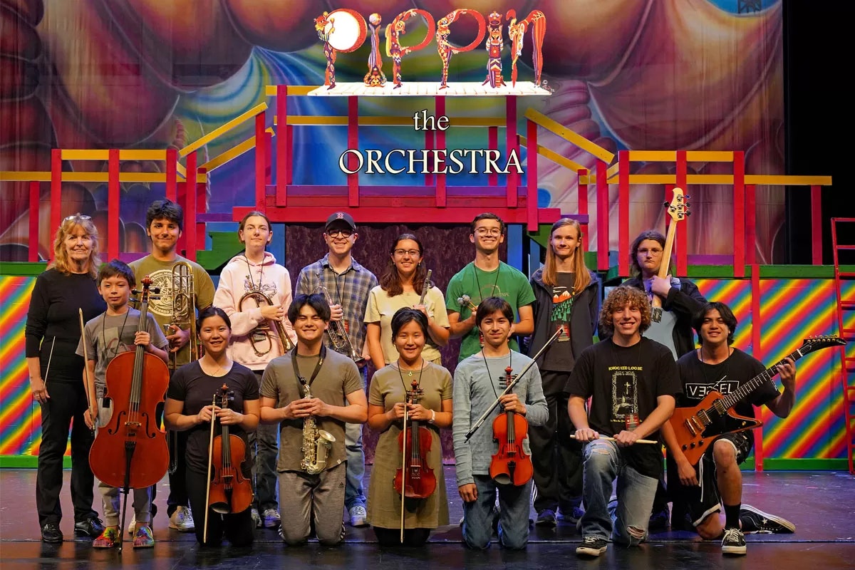 A group of musicians with various instruments pose in front of a colorful stage set for "Pippin: The Orchestra."