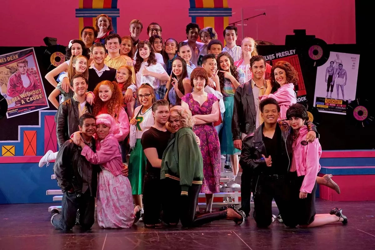 A group of people in colorful costumes posed on stage, resembling a theatrical production. Posters in the background.