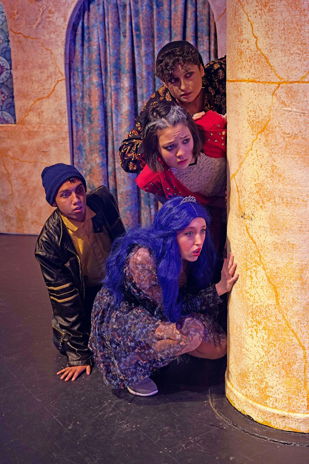 Four people in costume are peeking around a pillar on a stage.