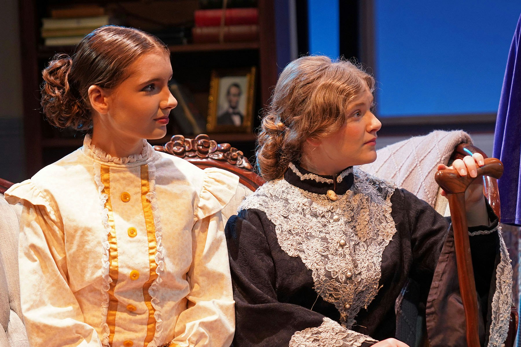 Two young women in vintage clothing are seated, engaged in a conversation, with a bookshelf and a framed picture in the background.